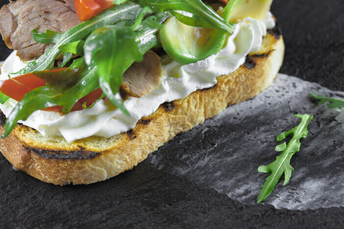 bruschetta with roast beef, cream sauce, avocado, tomato and arugula lies on a stone, mica stand, which stands on a wooden surface