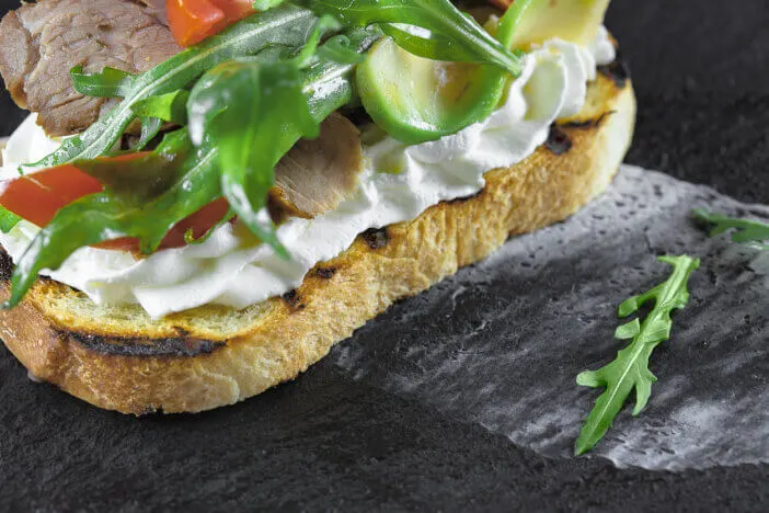 bruschetta with roast beef, cream sauce, avocado, tomato and arugula lies on a stone, mica stand, which stands on a wooden surface