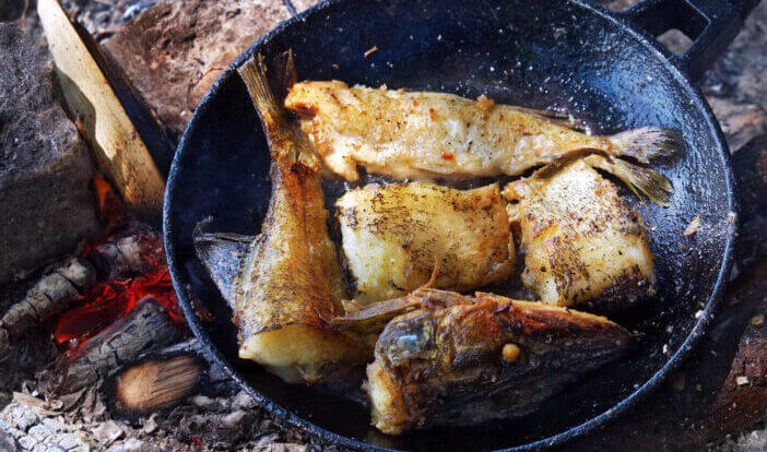 6+ Advantages of Cooking with a Cast Iron Skillet 1 Fried river fish in a cast iron skillet on hot ashes from the fire outdoors.
