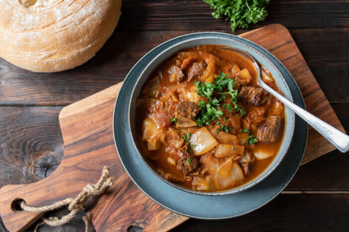 Homemade delicious brown beef stew with cubed meat, white cabbage and vegetables. Served in a bowl with fresh baked ciabatta bread on rustic and wooden table background. Top view
