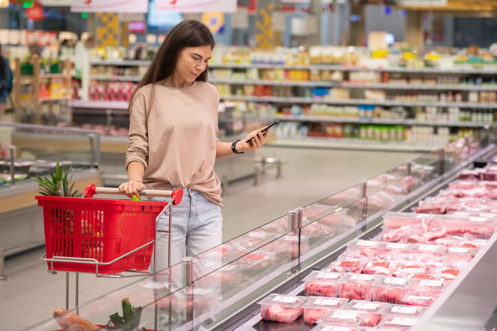 Young woman standing in aisle with shopping cart choosing fresh raw meat in modern supermarket