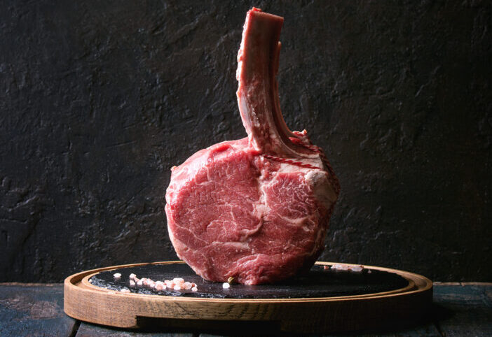 Raw uncooked black angus beef tomahawk steak on bone served with salt and pepper on round wooden slate cutting board over dark wooden plank table. Rustic style. Square image