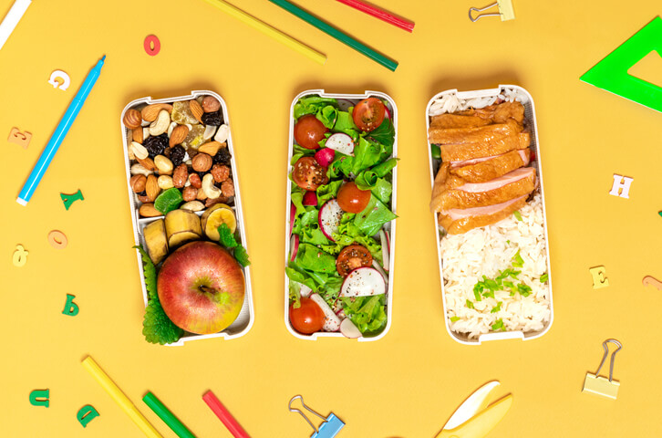 School lunch and stationery. Lunch boxes with chicken, rice, salad, fruits and nuts on an orange background, top view. Healthy balanced food for children in lunchboxes, flat lay.