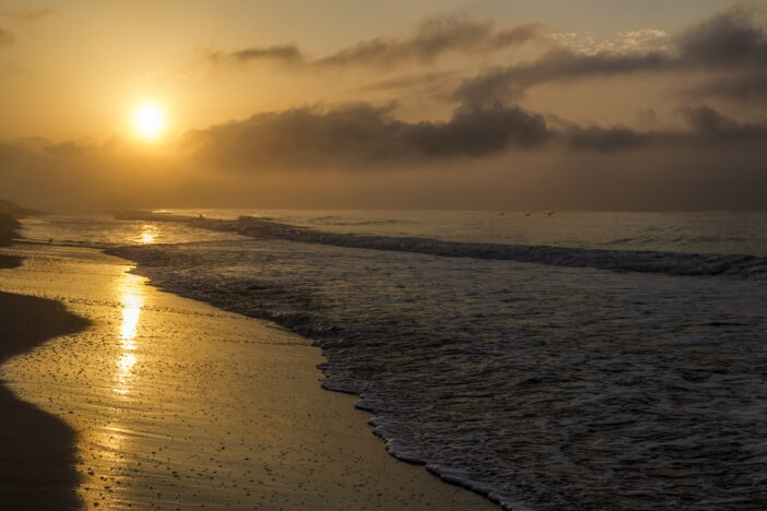 The golden morning light of sunrise shines on coastal Gulf Shores, Alabama with ocean waves gently breaking on the sandy beach and sea.