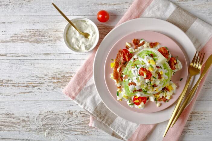 iceberg wedge salad topped with blue cheese dressing, crispy fried bacon, cherry tomatoes, crumbled hard-boiled eggs, chives on pink plates on a white wooden table, flat lay, free space, close-up