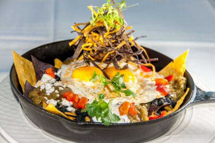 A delicious looking skillet contains a heaping portion of Huevos Rancheros.  Sunny up eggs, tortilla chips, fresh peppers and salsa are all visible in this yummy plate!