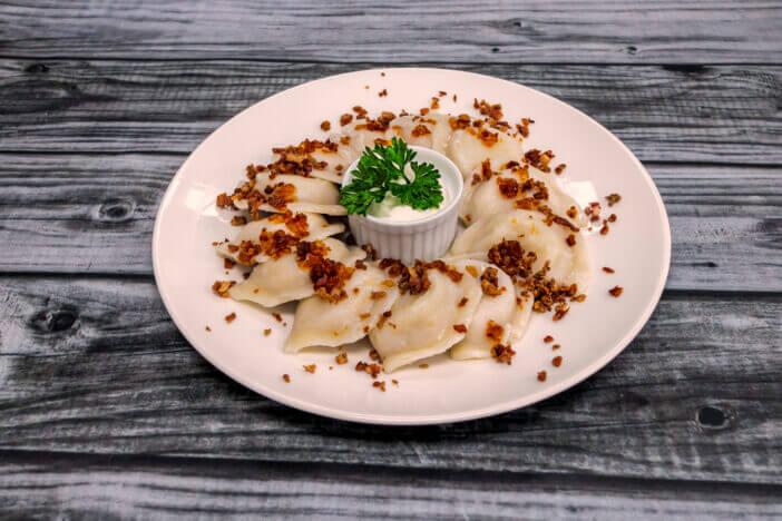 A large, round, white plate with a dish of sour cream in the center and perogies decorated around it. On a dark wood table shot at a 45 degree angle.