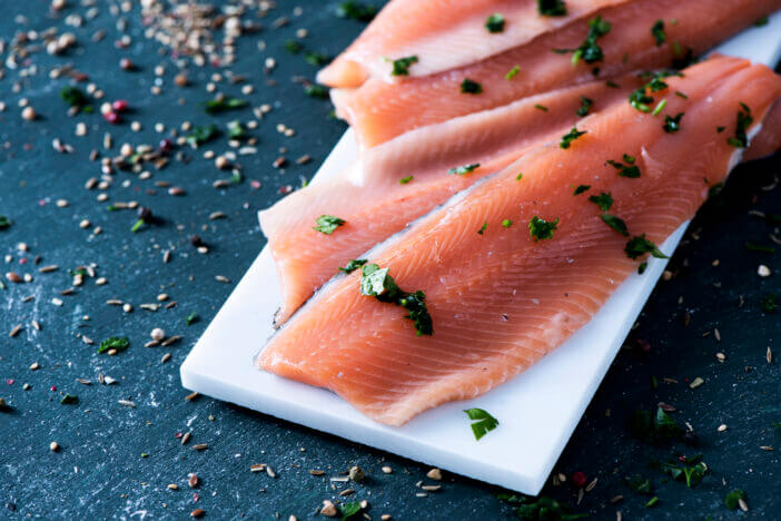 closeup of some slices of raw trout on a white marble tray, on a dark rustic surface sprinkled with herbs and spices