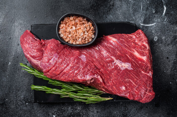 Raw American Tri Tip beef steak on marble board. Black background. Top view.