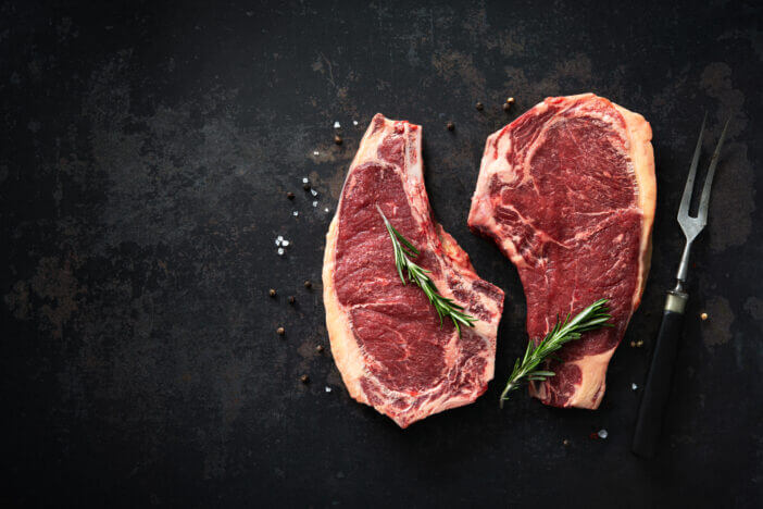 Two raw dry aged beef rib steaks (cote de boeuf) with rosemary, pepper and salt on dark background