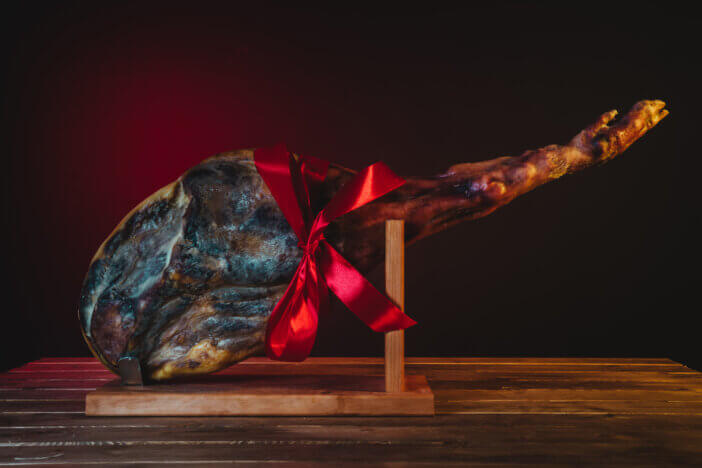 11 Special Occasion Meat Gifts For This Holiday Season 7 spanish ham placed in ham holder on a wooden table isolated with red and black background with a red gift bow of christmas